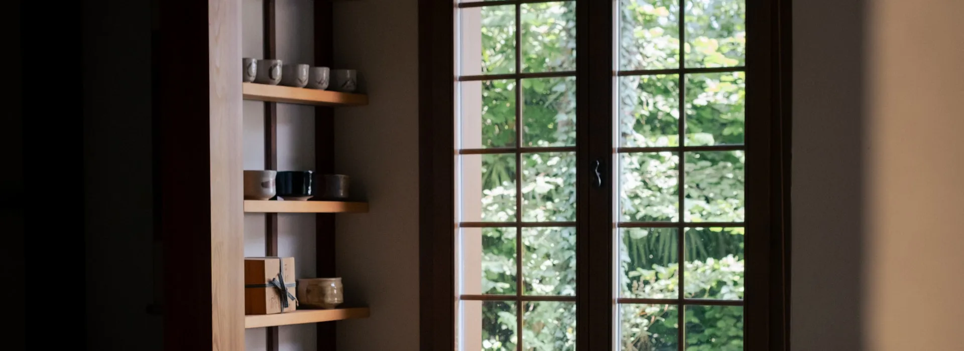Handmade chawan tea bowls displayed on a wooden shelf beside a window looking onto a green garden