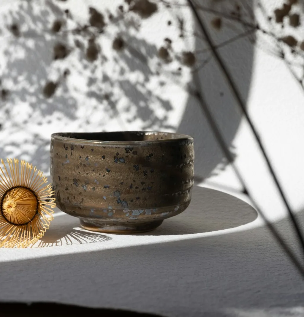A speckled glazed chawan and the tip of a bamboo chasen whisk on white textured paper, with a delicate branch shadow falling across the scene