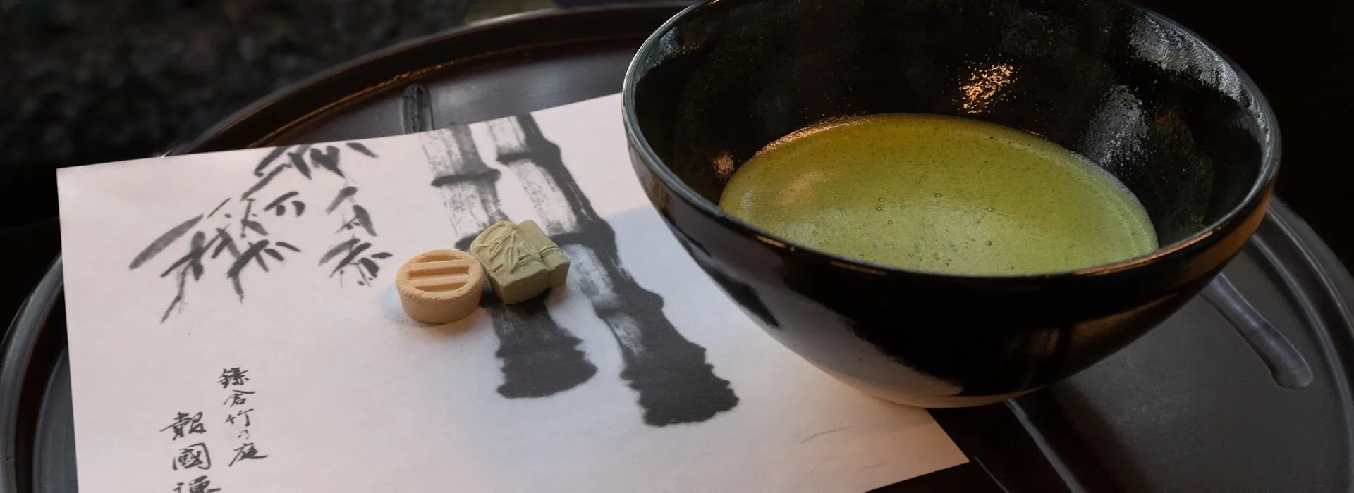 A single bowl of matcha and two wagashi sweets on a lacquered tray beside a hand-painted bamboo motif, a quiet solitary tea setting