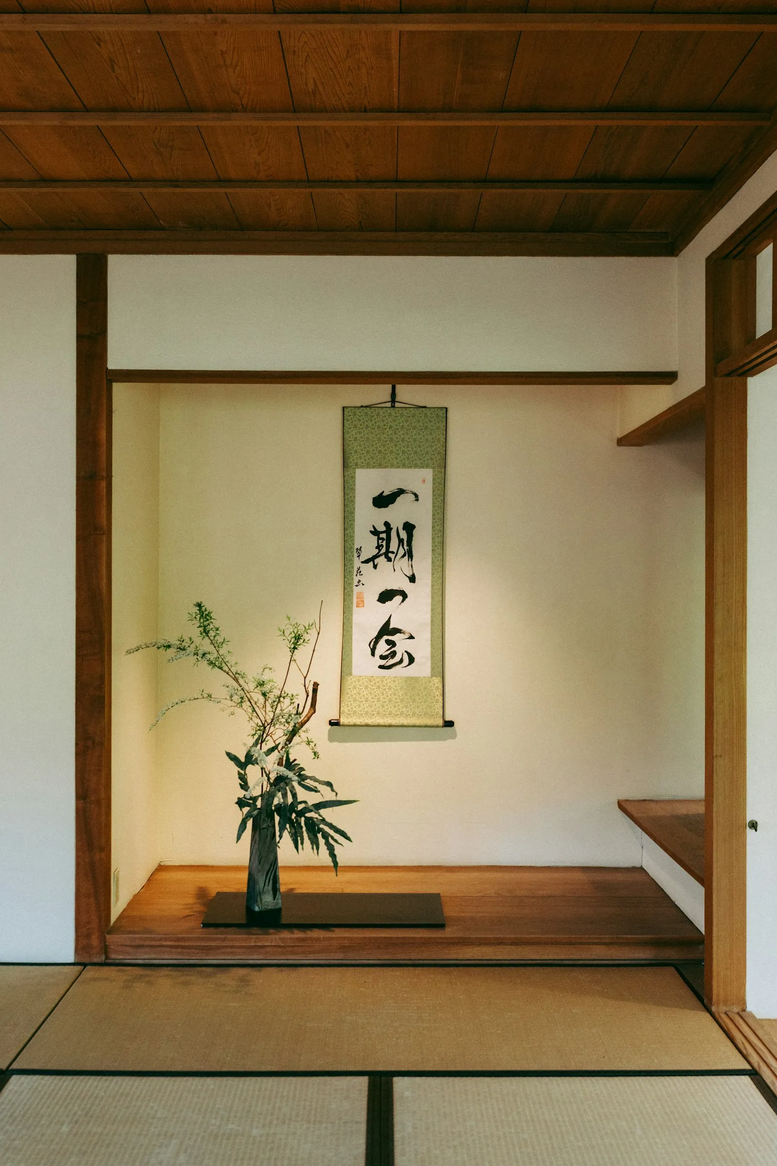 A tokonoma alcove with a hanging scroll reading ichi-go ichi-e beside an ikebana flower arrangement, in a traditional tatami tea room