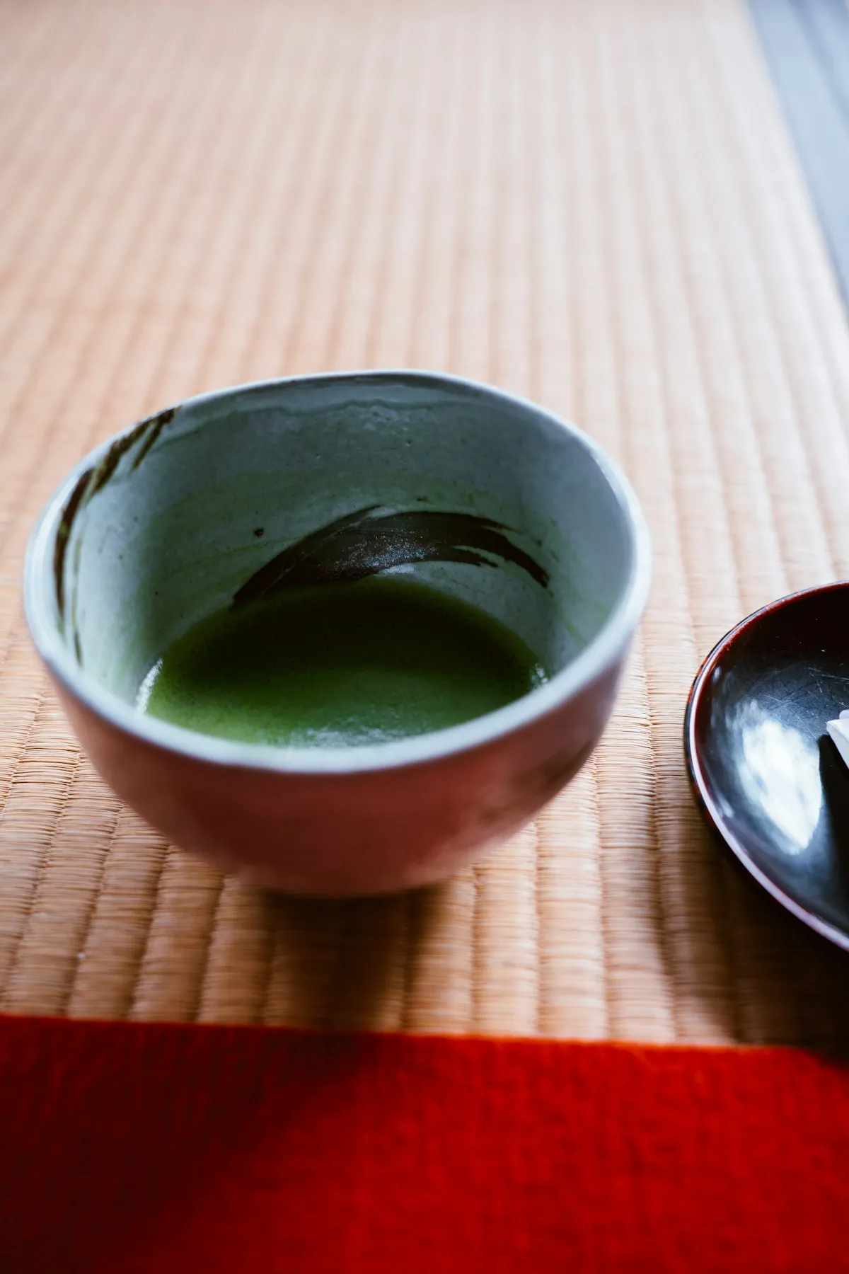 A glazed chawan with brushed markings holds freshly whisked matcha, set on a tatami mat beside a black lacquer dish