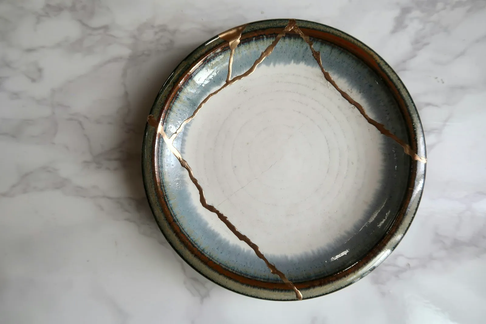 A ceramic plate on a marble surface, cracked and rejoined with visible gold kintsugi repair lines