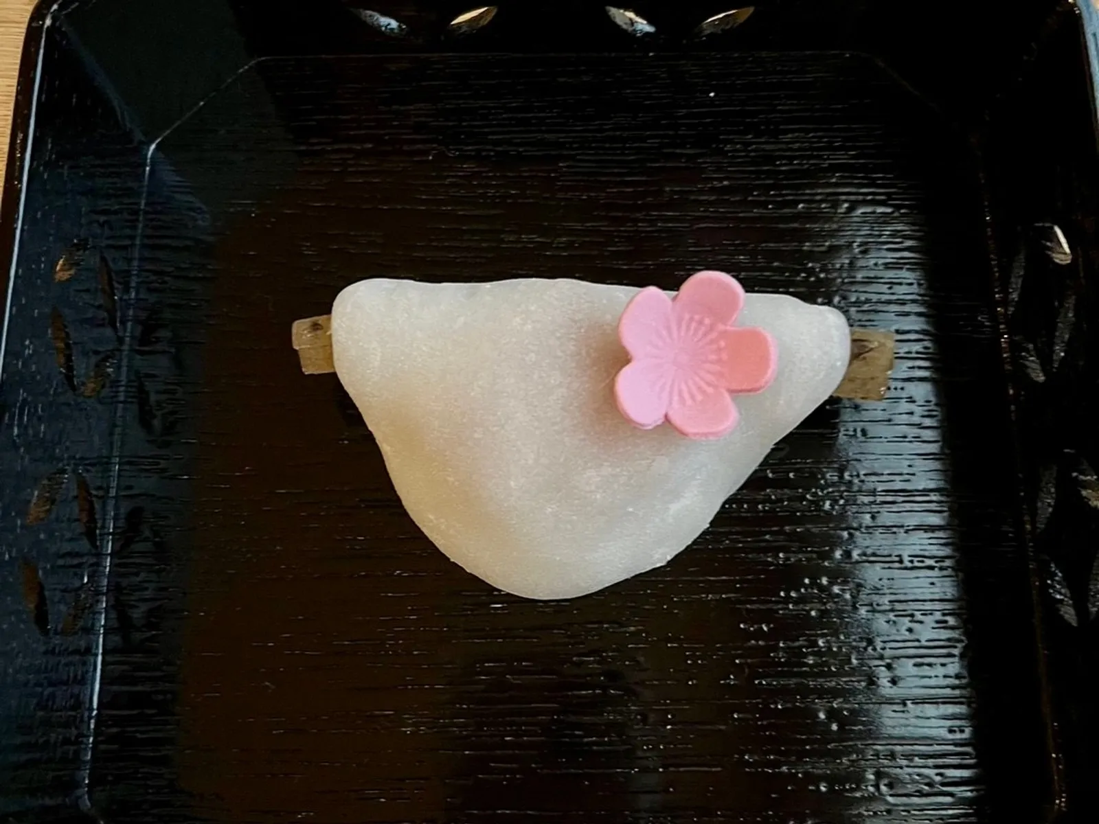 A white mochi wagashi with a pink sakura decoration on a black lacquer tray, with a kuromoji pick